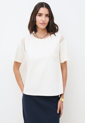 Femme aux cheveux châtains mi-longs portant un haut blanc à manches courtes avec des détails en dentelle, une jupe bleu marine, un collier en chaîne dorée et un large bracelet doré.