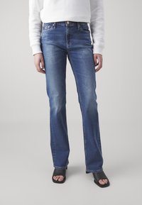 Jeans de mezclilla azul de campana con un acabado desvanecido, diseño de cinco bolsillos y botón de latón. Llevados con calzado negro de dedo al descubierto.