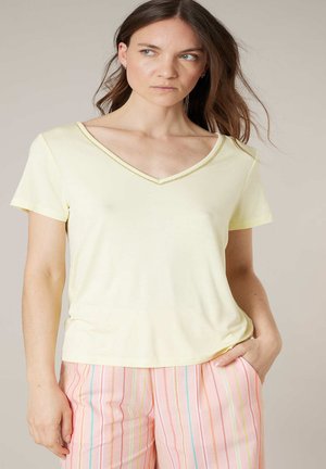Femme aux longs cheveux bruns portant un t-shirt jaune pâle en V et un pantalon à rayures roses pastel, une main dans la poche, regardant sur le côté.