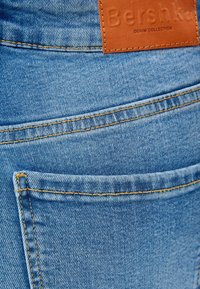 Světlé modré denimové džíny s koženou visačkou označenou "Bershka Denim Collection." Obsahují viditelné švy a design zadní kapsy.