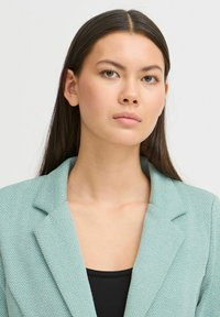 Lichtblauwe blazer met visgraatpatroon, pieklapels en textuurstof, met daaronder een zwarte top. Directe frontale weergave.