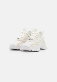 Sneaker bianche e beige pallido con una parte superiore in rete, sovrapposizioni testurizzate, suola spessa e design con lacci, completo di un dettaglio del logo sulla linguetta.