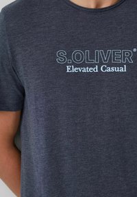 Dunkelgraues T-Shirt mit kurzen Ärmeln und hellblauem Text "S. OLIVER Elevated Casual" auf der Brust, getragen von einer Person.