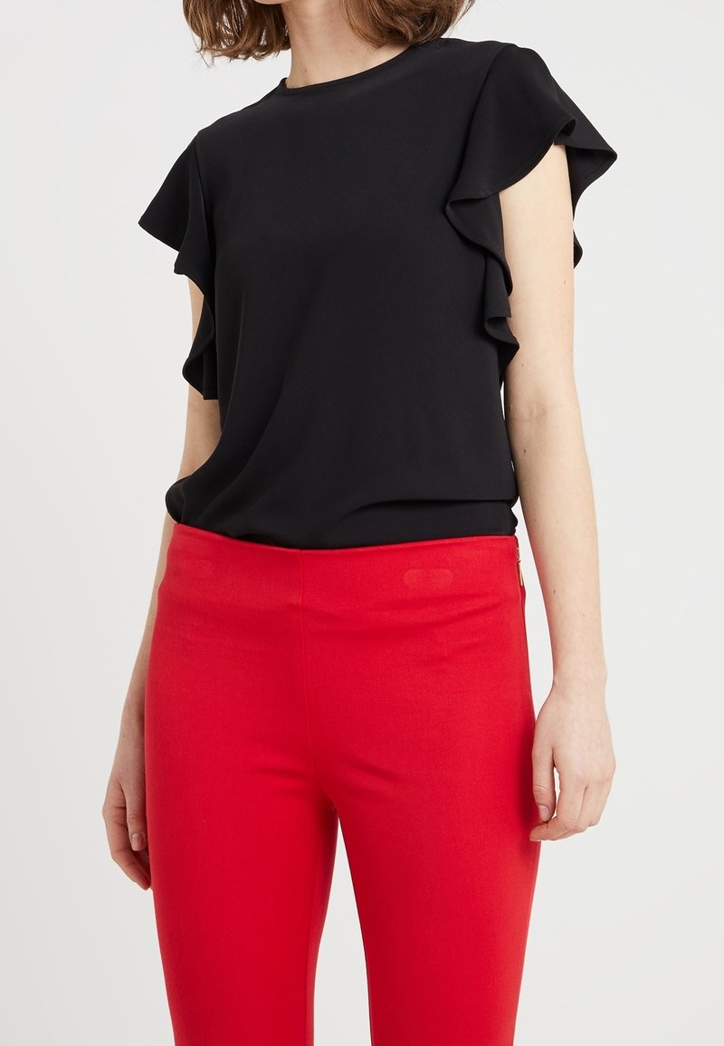 Kvinna i en svart kortärmad blus med volangärmar och högmidjade röda tajta byxor, stående framför en enfärgad bakgrund.