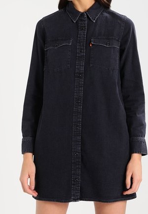 Kvinde iført en langærmet sort denimkjole med knapper foran og to brystlommer med klapper.