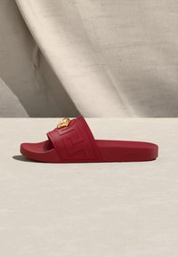 Sandale slide rouge avec une bride texturée présentant un motif clé grec et un emblème tête de lion doré sur le dessus.