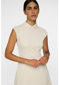 Nainen, jolla on tummat hiukset taakse sidottuna, pukeutuneena hihattomaan beigeen mekkoon, jossa on hienovarainen tekstuurikuvio ja pieni kaulus, kädet ristissä edessä.