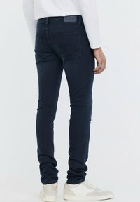 Jean skinny bleu foncé avec une texture lisse, cinq poches et un patch en cuir à la taille ; associé à des baskets blanches.