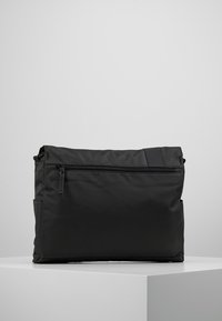 Svart nylon budväska med en texturerad yta, en frontficka med dragkedja och en strömlinjeformad design. Inkluderar en sidoficka för extra förvaring.