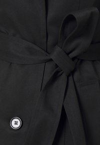 Cinturón de tela negro atado en un nudo en una prenda con costuras visibles y un botón blanco y negro cerca de la parte inferior izquierda.