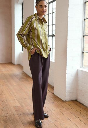 Grünes gestreiftes Button-up-Hemd mit einer entspannten Passform, kombiniert mit dunkelbraunen weiten Hosen und schwarzen Slippern. Hintergrund aus Naturholzboden.