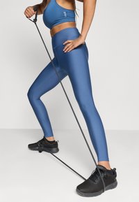 Μπλε αθλητικά κολάν με λεία υφή, ψηλή μέση και συνδυασμένο αθλητικό σουτιέν; σε συνδυασμό με μαύρα αθλητικά παπούτσια και λάστιχο αντίστασης.