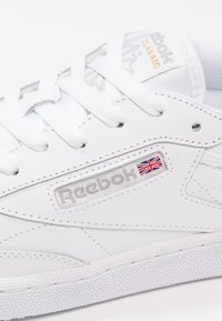 Detailní záběr na bílé tenisky Reebok Classic, zobrazeny šňůrky, šití, texturovaná podrážka a logo Reebok s vlajkou Union Jack.