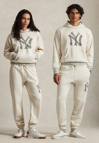 Lys beige hettegenser og joggebukser sett med en stor grå NY-logo, snøring i livet og ribbestrikkede mansjetter. Myk, teksturert stoff.