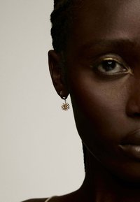 Pendiente de aro dorado con un diseño de explosión solar y un emblema de ojo en el centro; acabado pulido y elegante que resalta sobre un tono de piel oscuro.