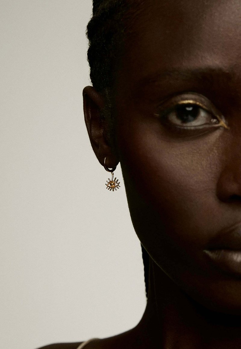Pendiente de aro dorado con un diseño de explosión solar y un emblema de ojo en el centro; acabado pulido y elegante que resalta sobre un tono de piel oscuro.