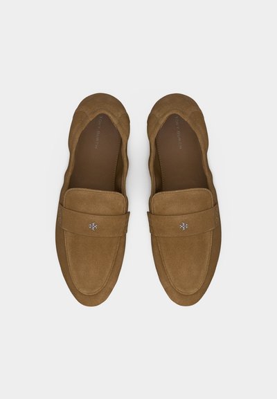Καφέ γυναικεία μοκασίνια από σουέτ με μικρό ασημένιο λογότυπο στις λουρίδες, στρογγυλεμένες μύτες και επενδεδυμένες σόλες σε λευκό φόντο.