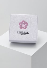 Balta teksturēta rotaslietu kaste ar rozā ziedu logotipu un melno tekstu "SENSORA JEWELRY" priekšā. Kvadrāta forma.