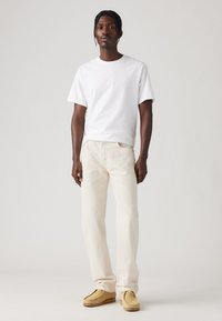 Hombre de pie vistiendo una camiseta blanca sencilla, jeans de color blanco roto y zapatos de gamuza beige, contra un fondo neutro.
