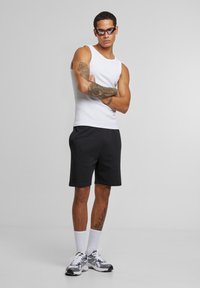 Man wearing red sunglasses, white tank top, black shorts, white socks, and sneakers, standing with arms crossed against plain background.