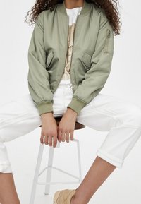 Personne assise sur un tabouret portant une veste bomber vert clair, un t-shirt graphique, un pantalon blanc retroussé et des baskets beiges.