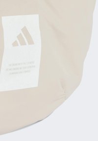 Bolso beige con una superficie suave y texturizada. Presenta un logotipo blanco y un panel de texto, con un diseño distintivo de tres franjas.