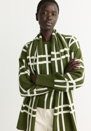 Woman with short hair wearing a green and white checkered sweater and matching scarf, arms crossed, standing against a plain background.