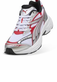 Baskets de sport blanches avec des accents argentés et rouges, tige en maille, semelle épaisse et semelle extérieure en caoutchouc noir. Équipées de lacets blancs et du logo Puma.