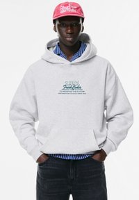 Felpa grigia chiara con tasca frontale, con testo blu che legge "1991 Fresh Codes" e una camicia a righe blu sotto.