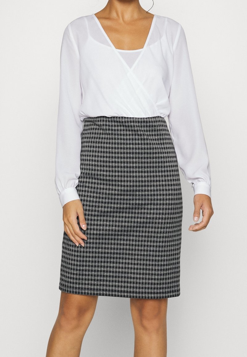 Femme portant un chemisier blanc à manches longues avec un effet cache-cœur, rentré dans une jupe taille haute mi-longue à carreaux noirs et gris.