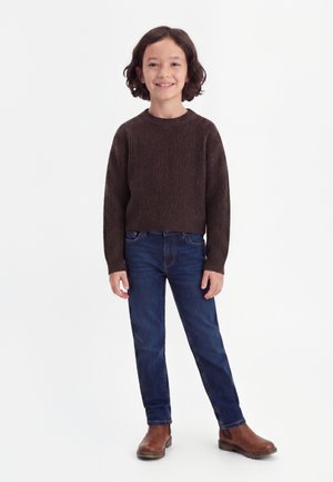 Copil cu păr întunecat până la umeri zâmbește, poartă un pulover maro tricotat, blugi albaștri închiși și ghete maro, stând în fața unui fundal alb.