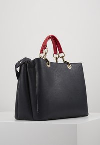 Czarna skórzana torebka o teksturowanym wykończeniu, z czerwonymi uchwytami, złotymi elementami i zorganizowaną konstrukcją z bocznymi fałdami.
