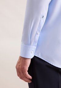 Camicia a maniche lunghe azzurro chiaro con polsini arrotondati e un bottone bianco, impreziosita da delicati ricami floreali sulle maniche. Tessuto con una texture morbida.