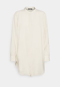 Lysebeige skjorte med lange ærmer, bandkrave, enkelt knaplukning og sidelommer for en afslappet pasform. Blødt stof med glat tekstur.