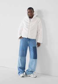 Witte nepbontjas met een hoge kraag en rits sluiting; gecombineerd met blauwe denim patchwork wijde broek en zilveren sneakers.