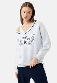 Vaaleanharmaa huppari, jossa on tummansiniset yksityiskohdat, ylipitkä malli ja kaareva kaula-aukko. Edessä on "LA NY" -teksti ja tähti-kuvio. Pehmeä kangas.