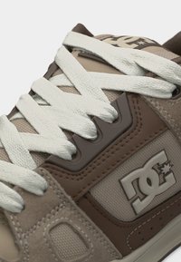 Κοντινό πλάνο μπεζ και καφέ αθλητικού παπουτσιού με λευκά κορδόνια και λογότυπο DC στην πλαϊνή επιφάνεια.