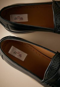 Zwarte leren instappers met een gestructureerd accent op de bovenkant, een glad interieur en een label met de tekst "Next Signature Italian Leather."