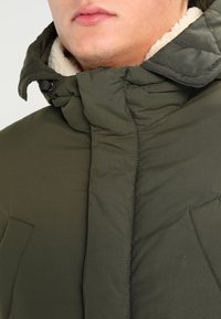 Oliwkowo-zielona kurtka puchowa z fakturowaną powierzchnią, wyposażona w przytulną, kremowobiałą podszewkę z polaru oraz wysoki kołnierz z ozdobnym przeszyciem.