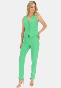 Zielona kamizelka z dekoltem w serek, z trzema guzikami i ostrym dołem, w zestawie z dopasowanymi spodniami i cielistymi sandałami na słupku.