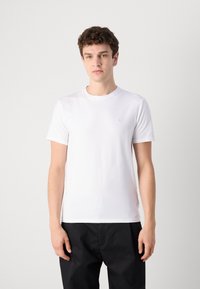 Witte katoenen t-shirt met een ronde hals en korte mouwen; voorzien van een klein grijs logo op de linkerborst; gladde textuur. Zwarte broek.