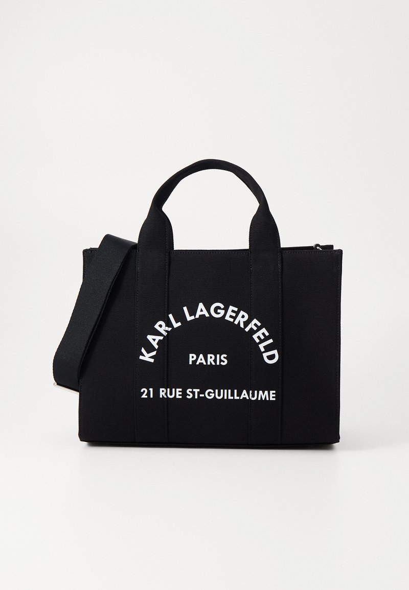 Svart totebag laget av slitesterk stoff. Har to topphåndtak, et justerbart reim, og stor hvit tekst som viser merke og adresse.