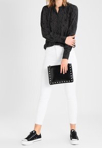 Blusa nera a maniche lunghe con motivi sottili, abbinata a jeans skinny bianchi e sneakers nere, mentre tiene una clutch nera con borchie.