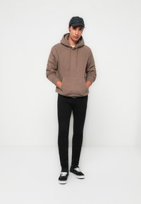 Man wearing brown fleece hoodie, black cap, black skinny jeans, and black sneakers with white laces, standing against a white background.