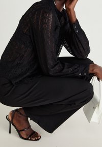 Blouse en dentelle noire à manches longues et col montant, associée à un pantalon noir et des talons noirs à brides. Tenant un petit sac à main blanc.