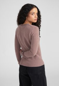 Suéter de punto en mauve claro con dobladillos y puños acanalados, cuello redondo, con una textura suave. La modelo tiene el cabello rizado y lleva pantalones negros.