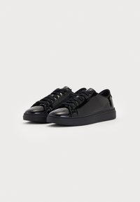 Mustad läikivad mustad madalad sneakers, millel on nöörid ja külgmine lukku, valgel taustal, kaldus paremale.