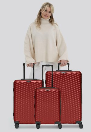 Trois valises rigides rouges avec un motif en chevrons, de tailles variées, poignées noires et roues. Placées sur un fond neutre.