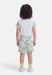 Παιδί όρθιο, με το πρόσωπο προς τα πίσω, φοράει λευκό πουκάμισο με μανίκια με σχέδιο και σορτς που ταιριάζουν με εκτυπώσεις ζώων της ζούγκλας και φύλλων, κίτρινες κάλτσες, άσπρα παπούτσια.