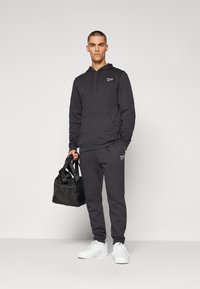 Haut à capuche en coton noir avec poche frontale et cordon de serage, associé à un pantalon de jogging assorti. Le mannequin tient un petit sac de sport noir. Baskets blanches.